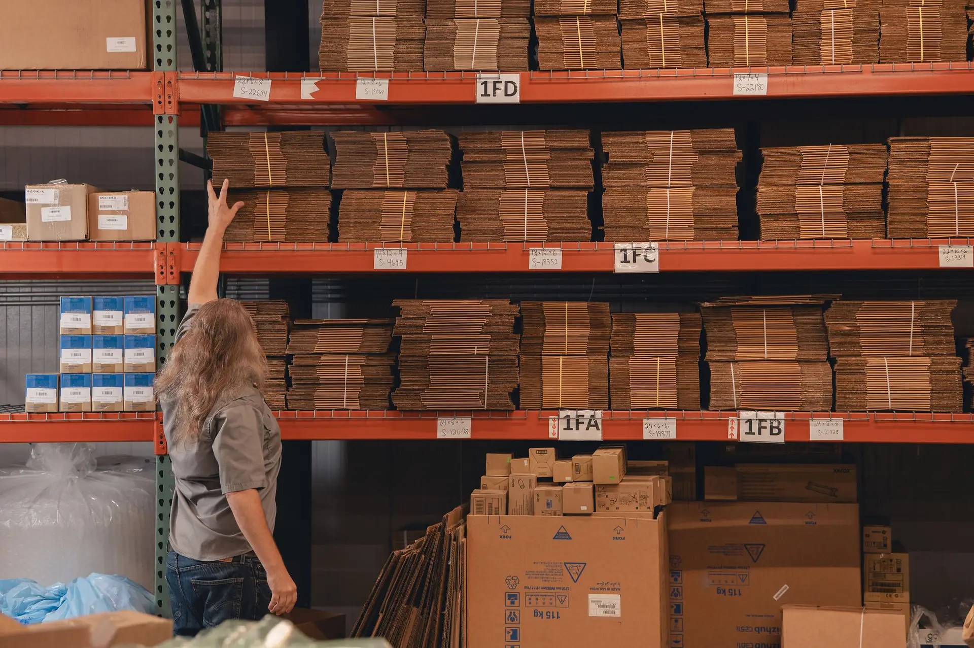 A person with long hair reaches up toward stacks of flat cardboard boxes stored on high metal shelves in a warehouse filled with various packaging materials and boxes.