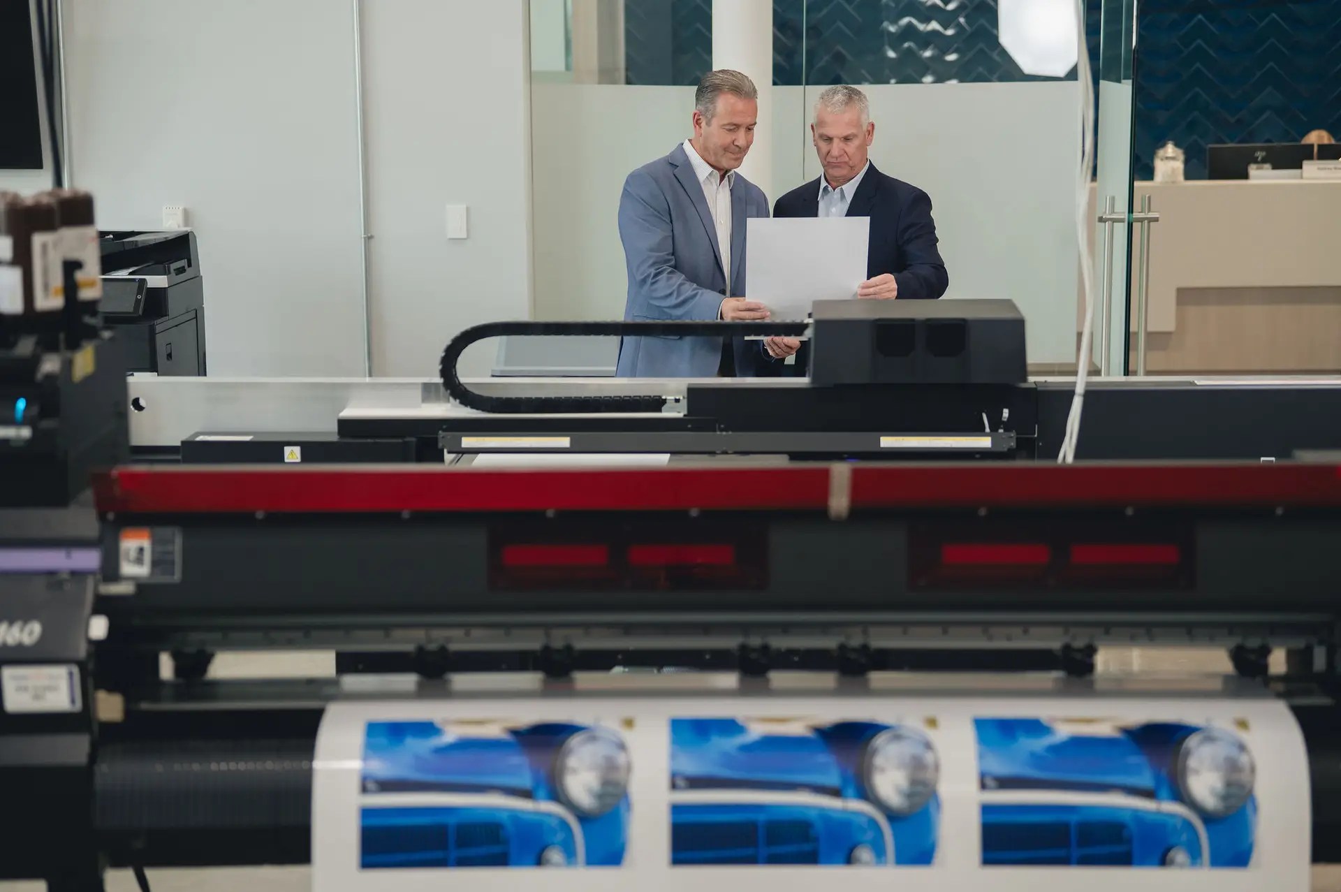 Two men in suits review a large printout in a modern print shop, standing behind a professional printer producing images of a blue car.