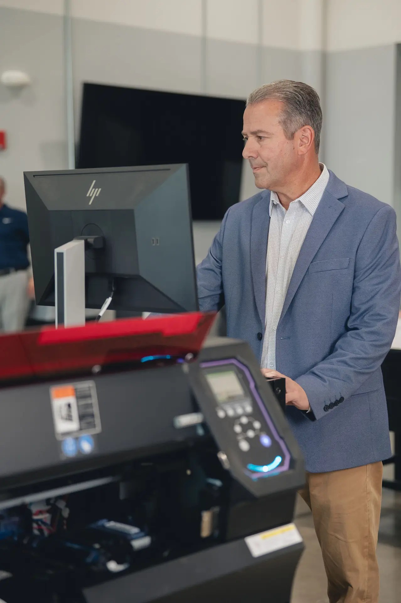 A man in a blue blazer and khaki pants stands at a desk, working on a computer. In the foreground, there is a large electronic device—possibly one of the latest printers and copiers Ohio offices rely on—in a tech workspace.