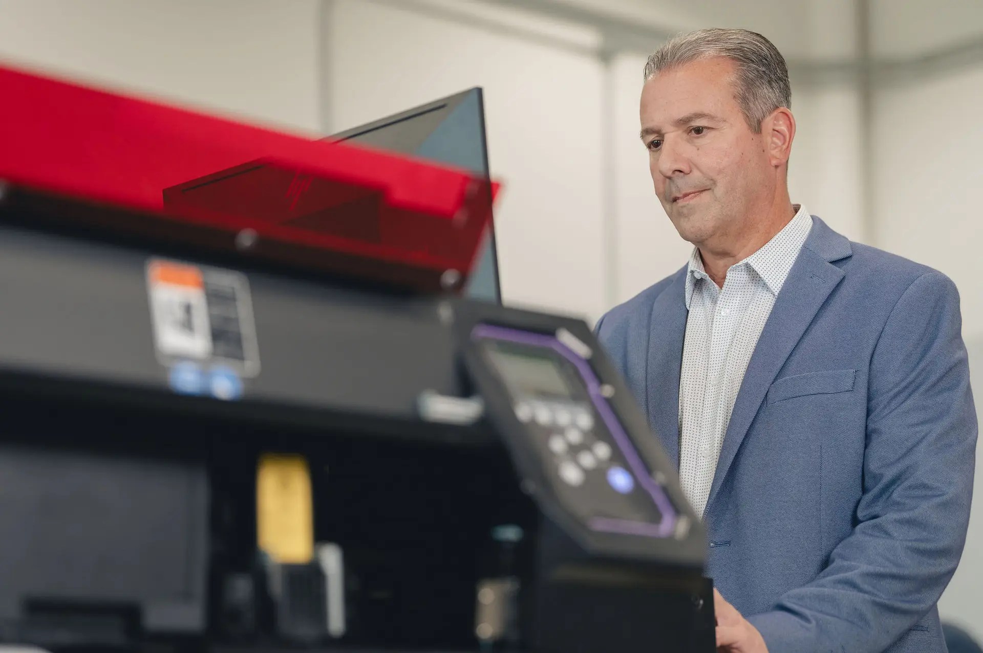 A man in a blue suit jacket operates a large black and red machine with electronic controls, showcasing advanced production print services Ohio in a modern, well-lit indoor setting.