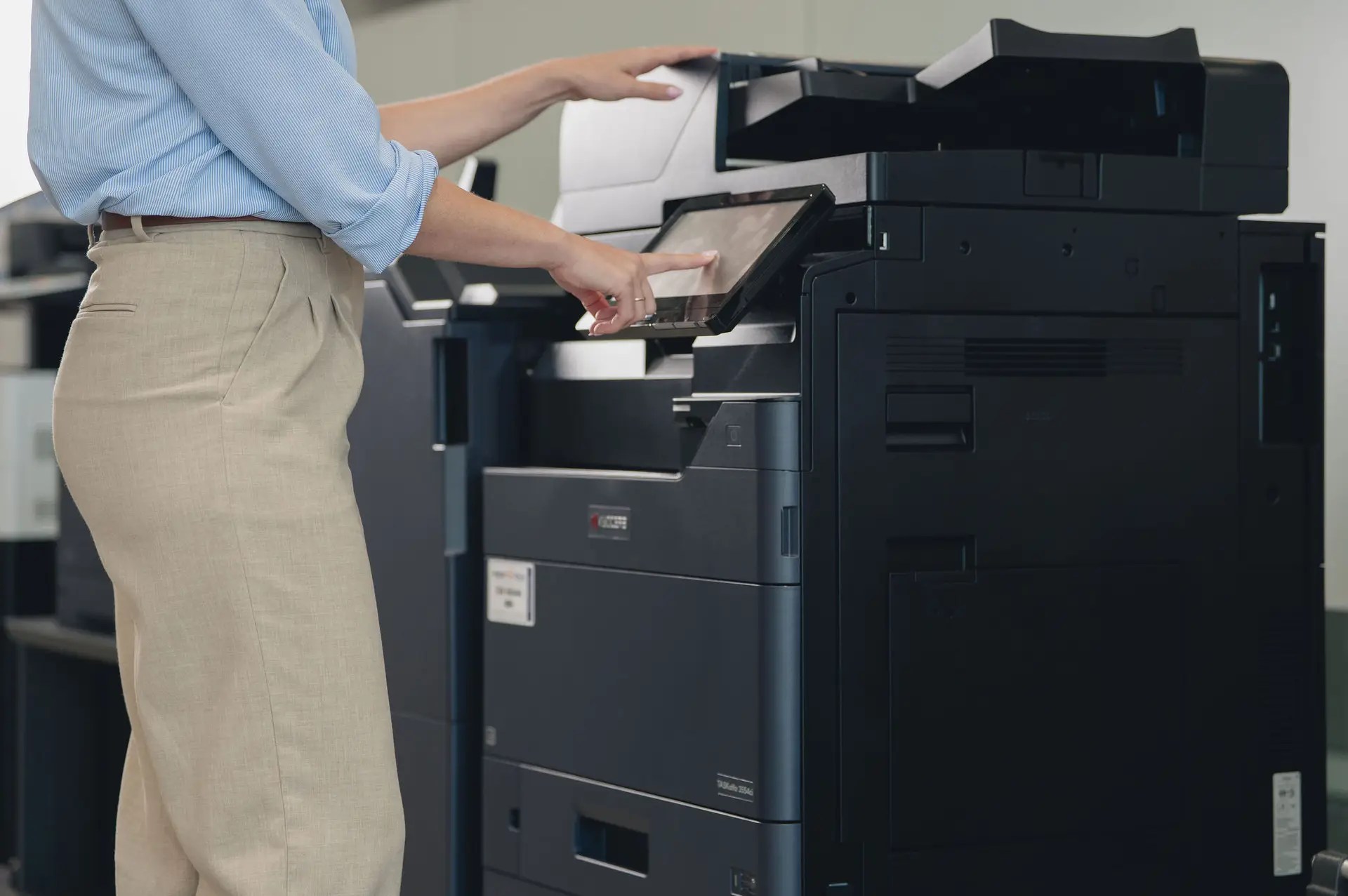 A person wearing beige pants and a light blue shirt is using the touchscreen control panel of a large black copier or printer in an office, showcasing modern printers and copiers Ohio businesses rely on for efficiency.