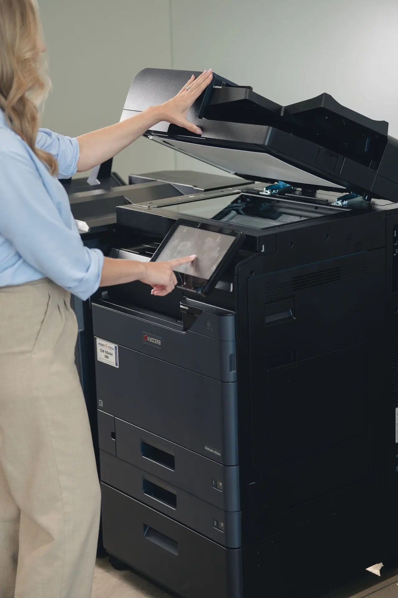 A person with long blond hair, wearing a blue shirt and beige pants, operates a large black office copier—an example of the equipment supported by managed print services Ohio—lifting the lid with one hand and using the touchscreen panel with the other.