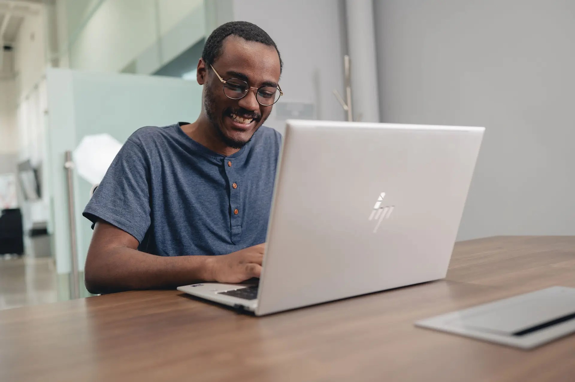 A smiling person wearing glasses and a blue shirt sits at a wooden desk, typing on a silver HP laptop in a modern, well-lit office space, focusing on Managed IT and Network Services.
