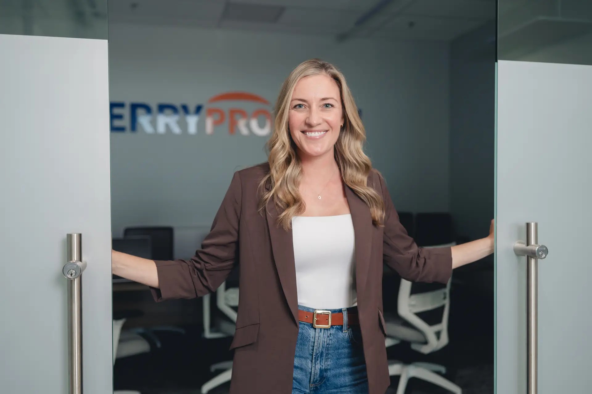 A smiling woman with long blonde hair, wearing a brown blazer, white top, and jeans, stands holding open glass doors in a modern office with white chairs in the background.