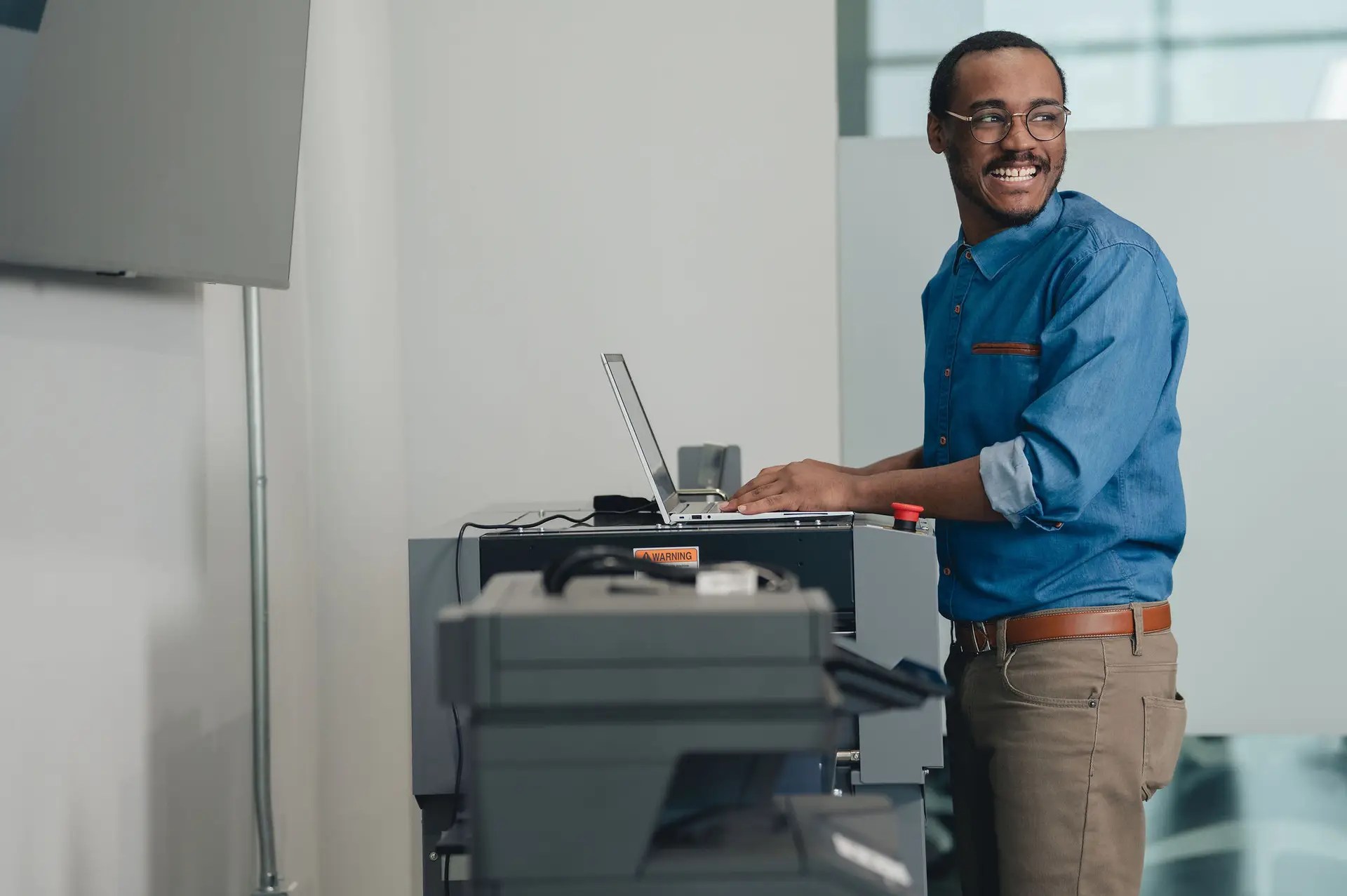 A smiling man in a blue shirt and glasses stands beside a large office printer, typing on a laptop in a modern, bright workspace.