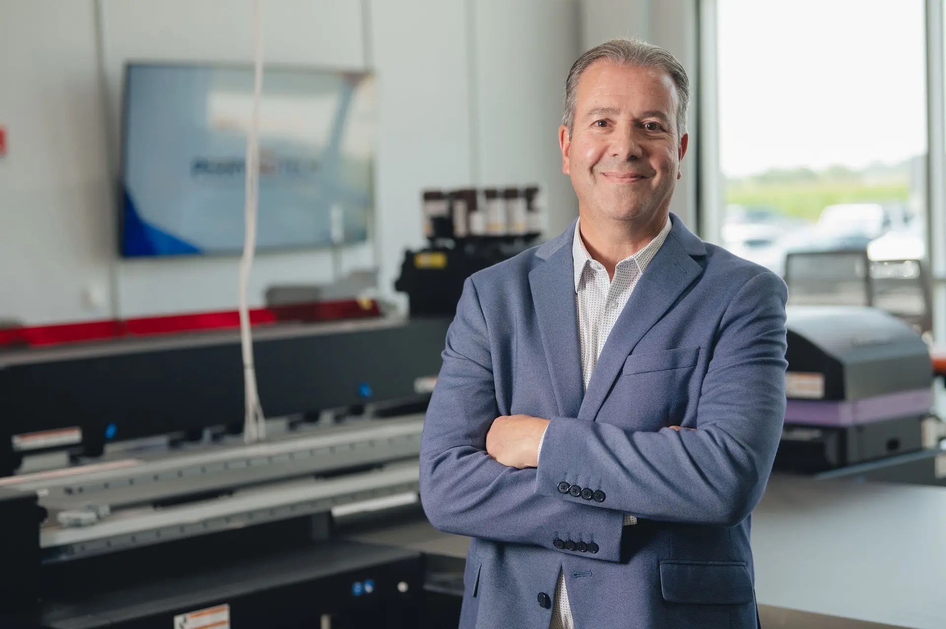 A man in a blue suit jacket stands with arms crossed and smiles in a modern office or printing facility, representing production print services Ohio, with large equipment and a window in the background.