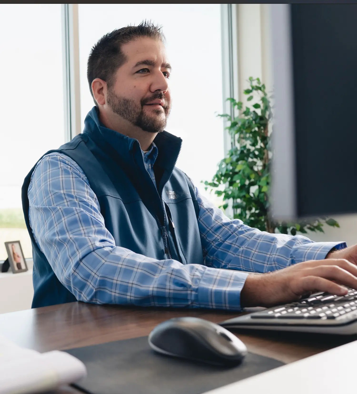 A man with short dark hair and a trimmed beard, wearing a blue plaid shirt and vest, sits at a desk typing on a keyboard in a bright office with large windows—a typical scene in document management Ohio.
