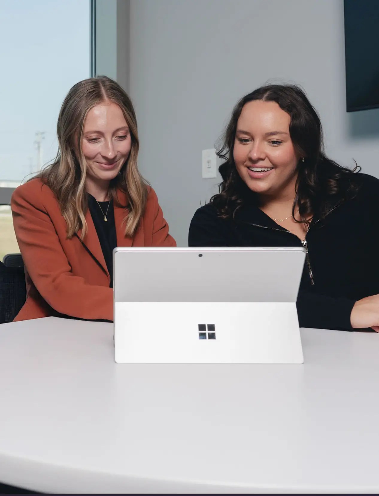 Two women sit at a white table, smiling and looking at a Microsoft Surface laptop together in a modern, bright office setting.