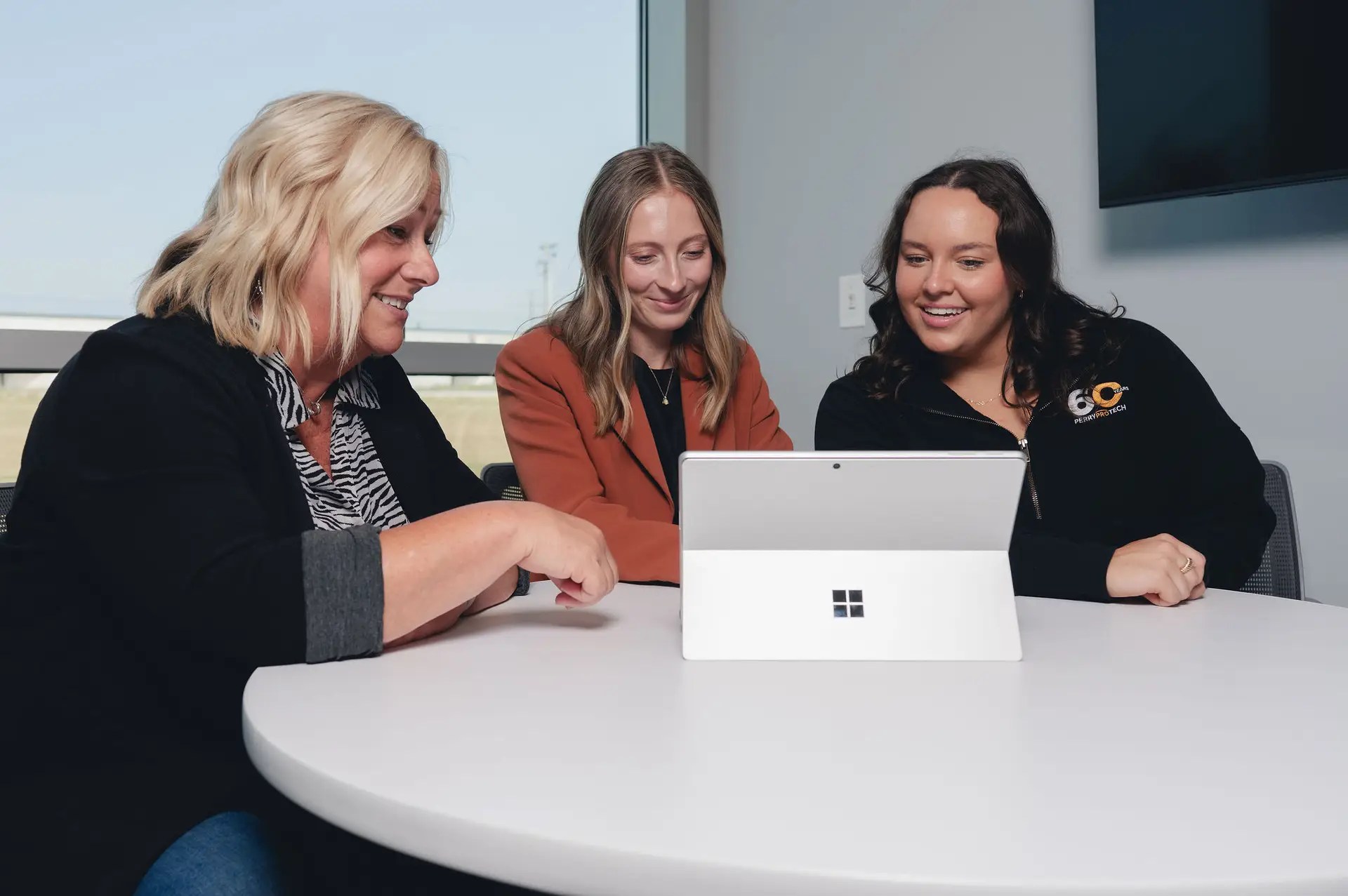 Three women sit at a round table, smiling and looking at a Microsoft Surface tablet together in a bright, modern Ohio office, showcasing the power of business technology with a large window in the background.