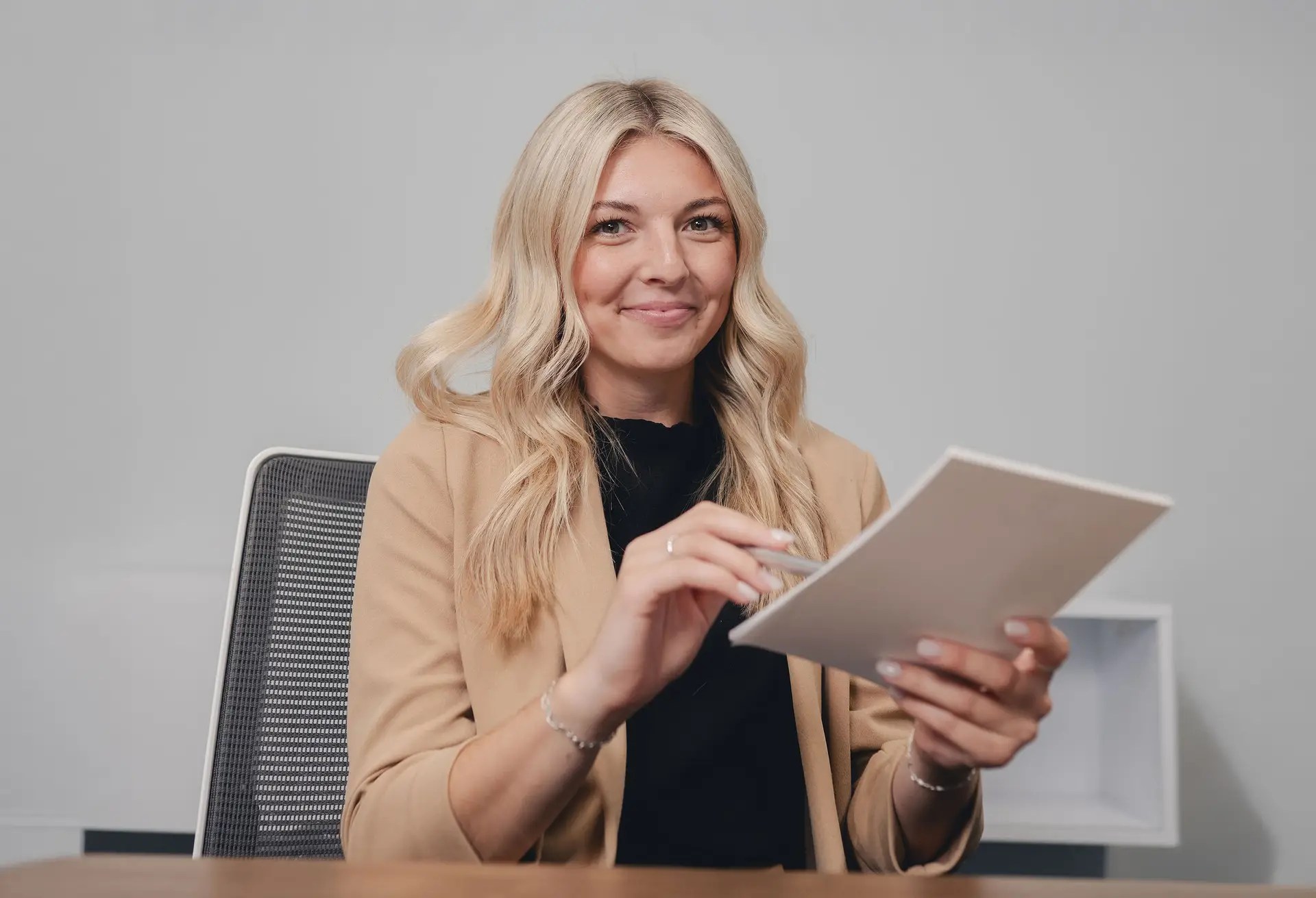 A woman with long blonde hair, wearing a tan blazer and black top, sits at a desk in Ohio, smiling and holding a digital tablet in her hands. The plain, minimalistic background highlights the integration of business technology.