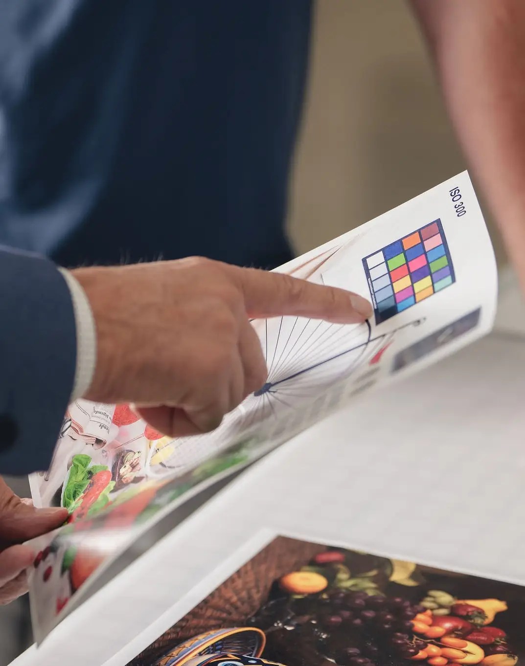 A person points to a color calibration chart printed on a sheet of paper, showcasing images of fruits and vegetables—an example of production print services Ohio—with another individual partially visible in the background.