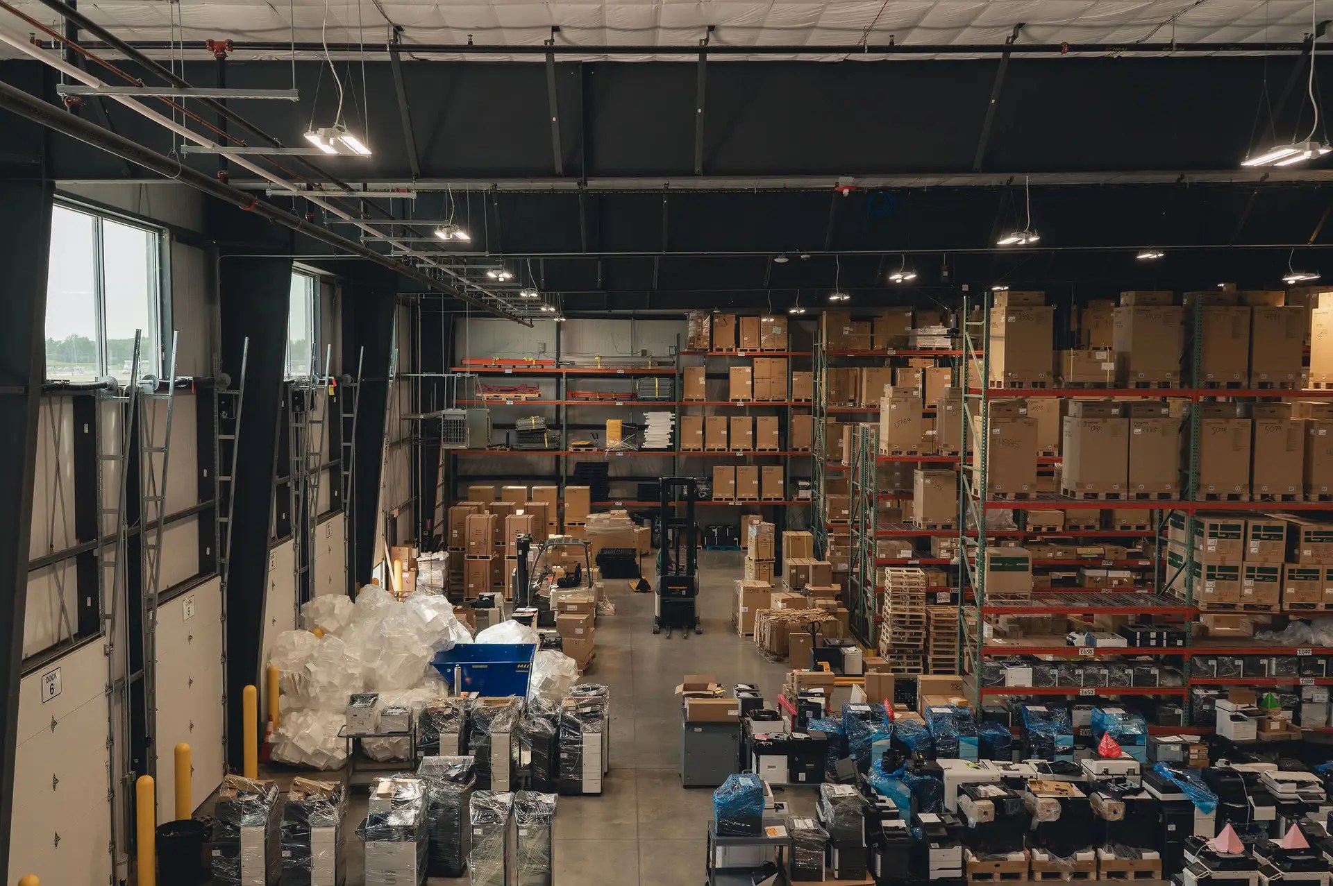 A large warehouse interior with high shelves filled with cardboard boxes and pallets. In the foreground, sorted items and bags are organized on tables, with people working among the storage racks and packing materials.