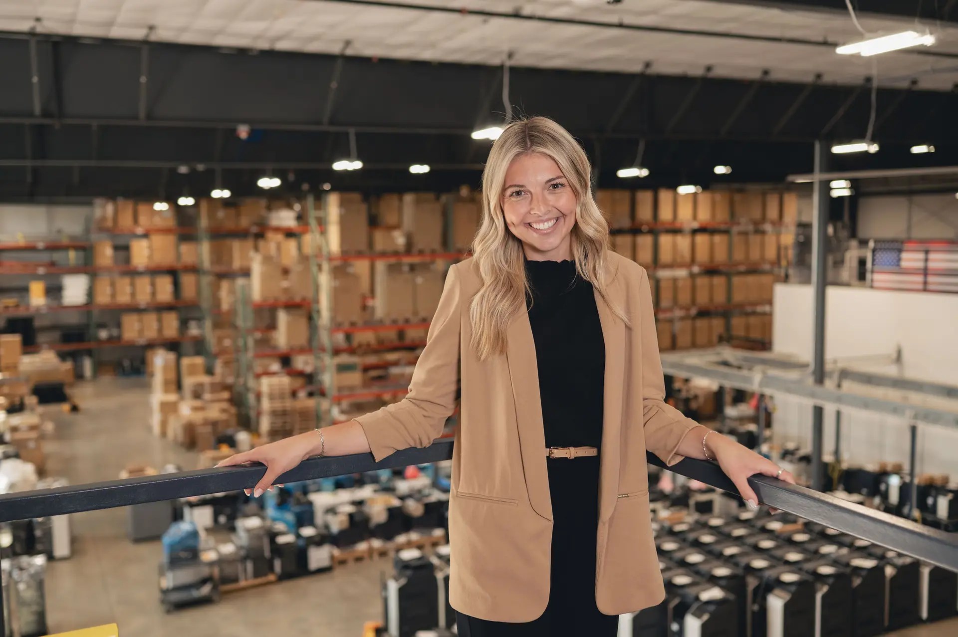 A smiling woman in a tan blazer stands on a balcony inside a large warehouse filled with boxes and packages. This Ohio business showcases efficient technology, with high ceilings and an American flag displayed inside.