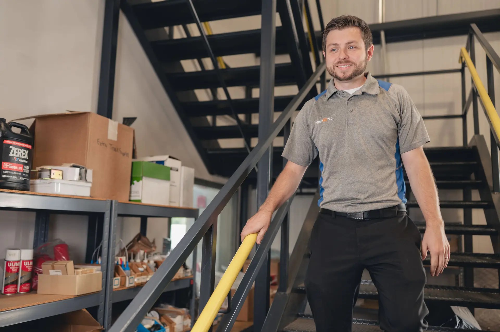 A man in a gray polo shirt and black pants smiles while walking down a metal staircase, holding the yellow handrail. Shelves with boxes and supplies are visible in the background.