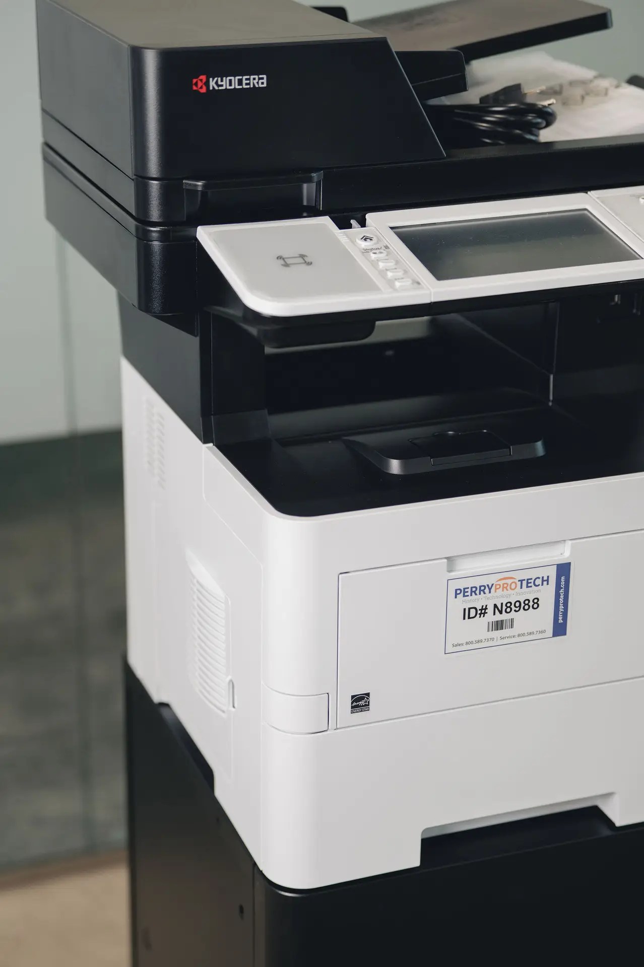 A close-up view of a Kyocera multifunction printer and copier, popular among printers and copiers Ohio users, featuring a display screen, keypad, and a Perry ProTech ID label on the front.