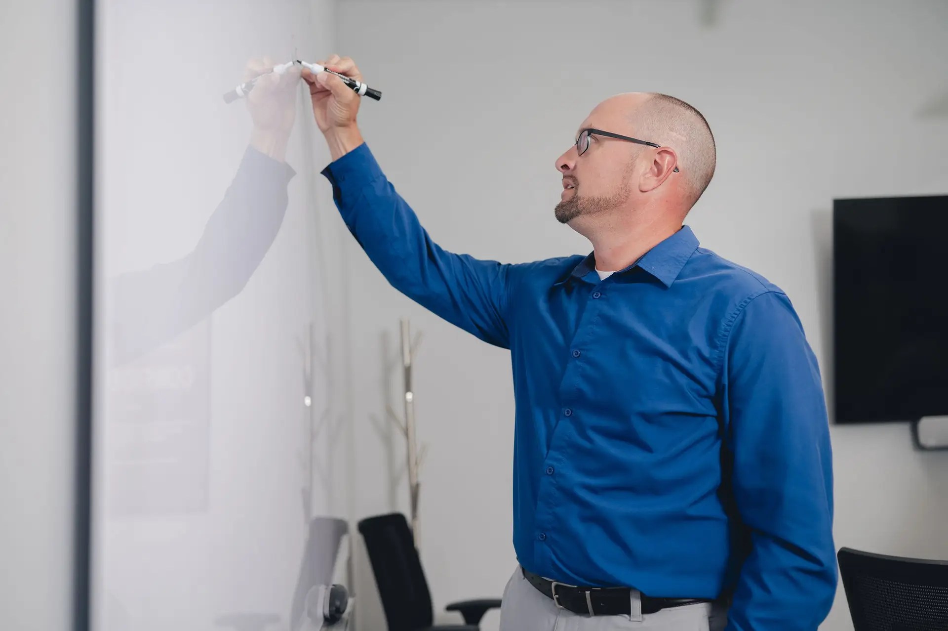 A man in a blue shirt and glasses writes on a whiteboard with a marker in a modern office setting.