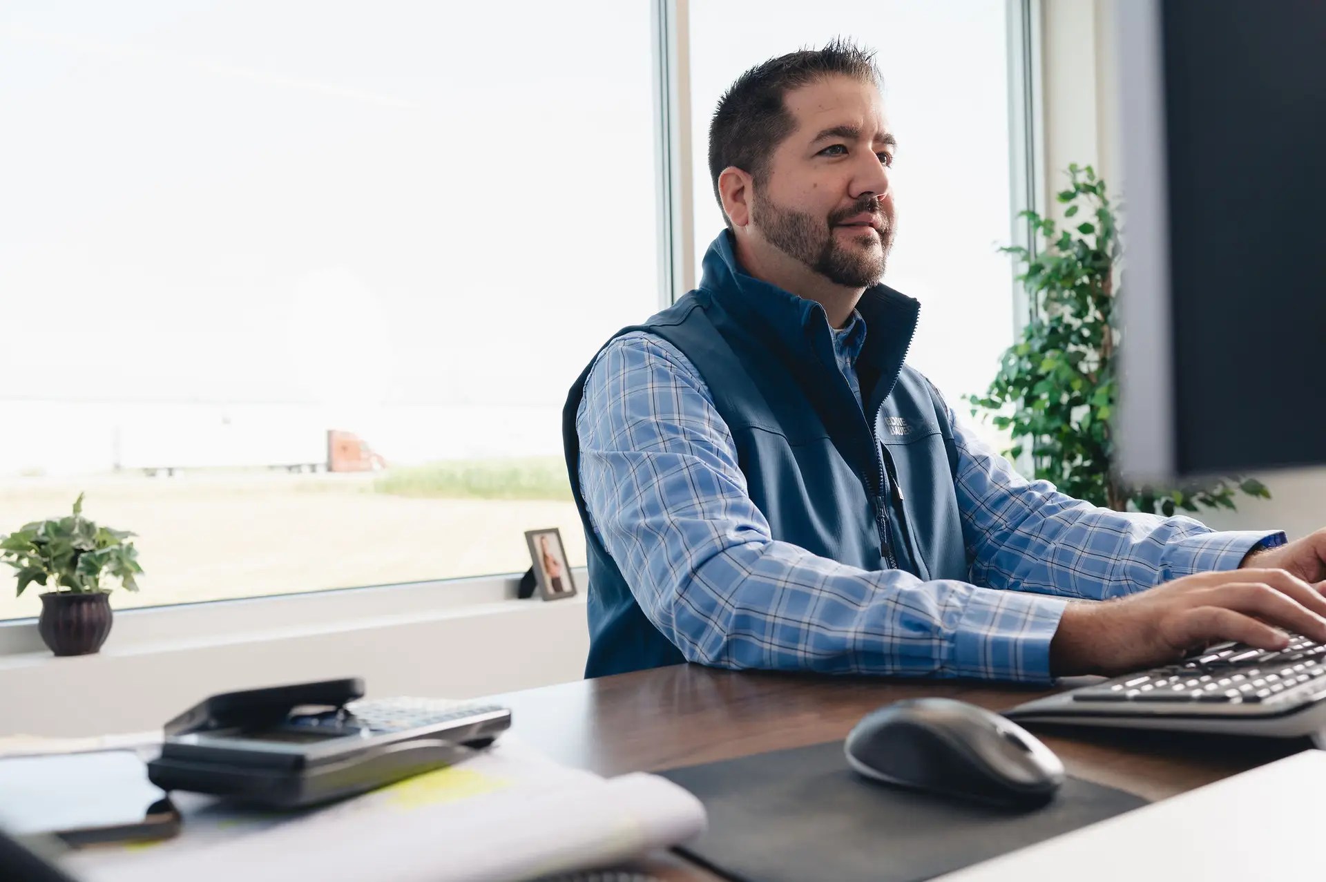 Man in a blue plaid shirt and vest sits at a desk working on a computer in a bright office, with paperwork, a plant, and calculator nearby. Large windows let in natural light.