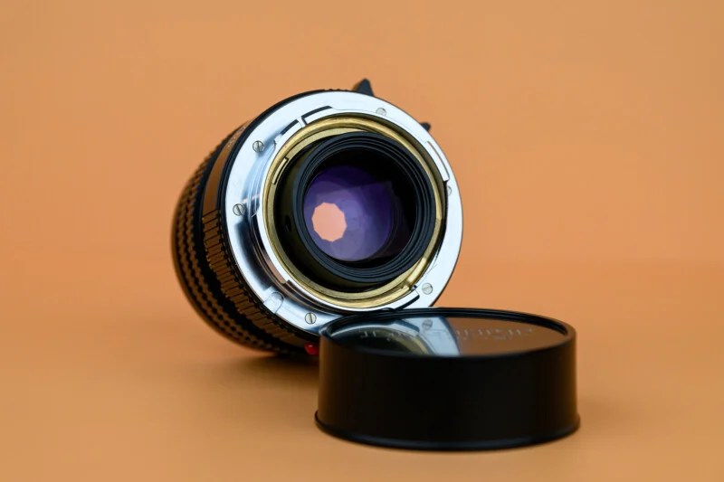 A camera lens lying on its side with the rear glass element and metal mount facing forward, next to a detached lens cap, all on an orange background.