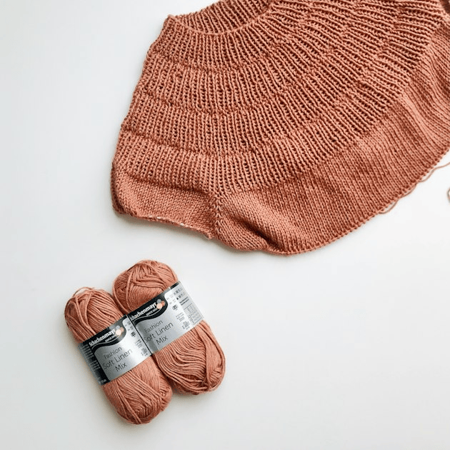 Rust-colored knitted piece beside a matching ball of yarn on a white surface.