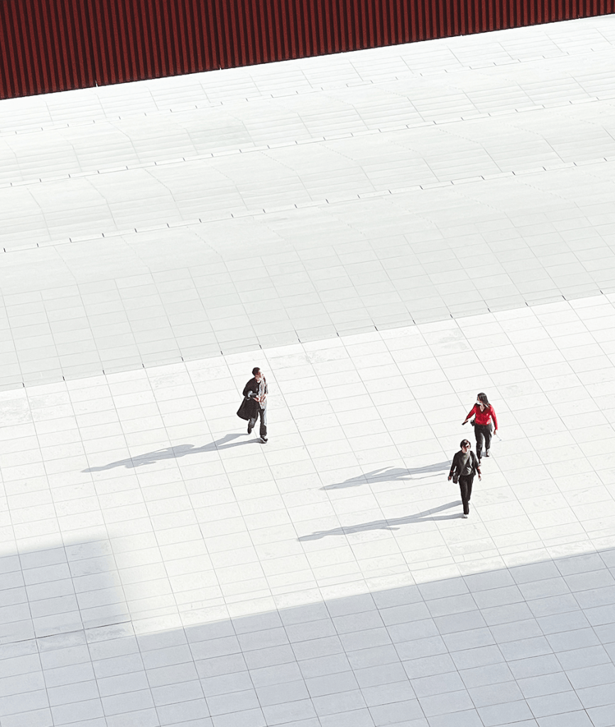 Una vista aérea de varias personas caminando sobre una superficie blanca, con sombras alargadas. Tres individuos se destacan, uno de ellos lleva una chaqueta negra, otro viste de rojo y el tercero tiene una mochila.