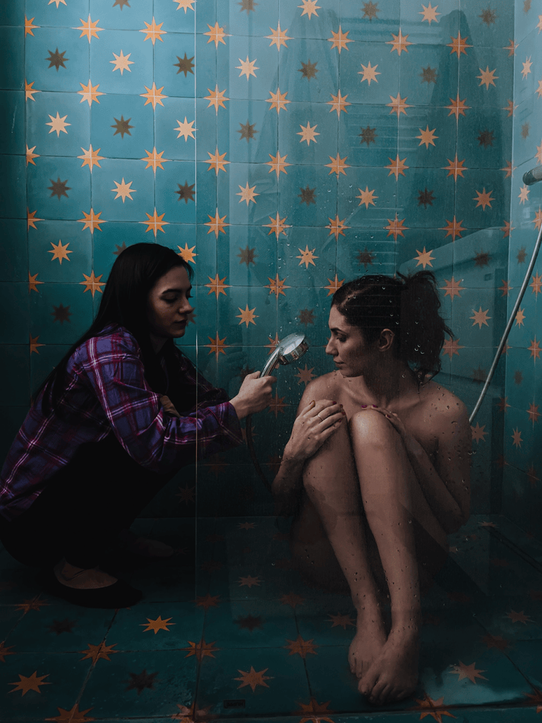 Una persona está tomando una fotografía de otra persona sentada en una ducha, rodeada de azulejos de color azul con estrellas naranjas.