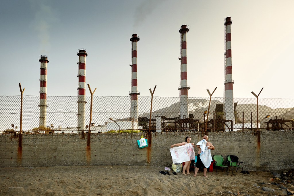 Dos personas en la playa, cubiertas con toallas, posan frente a chimeneas industriales al fondo. La escena muestra un contraste entre el ambiente costero y la infraestructura industrial.