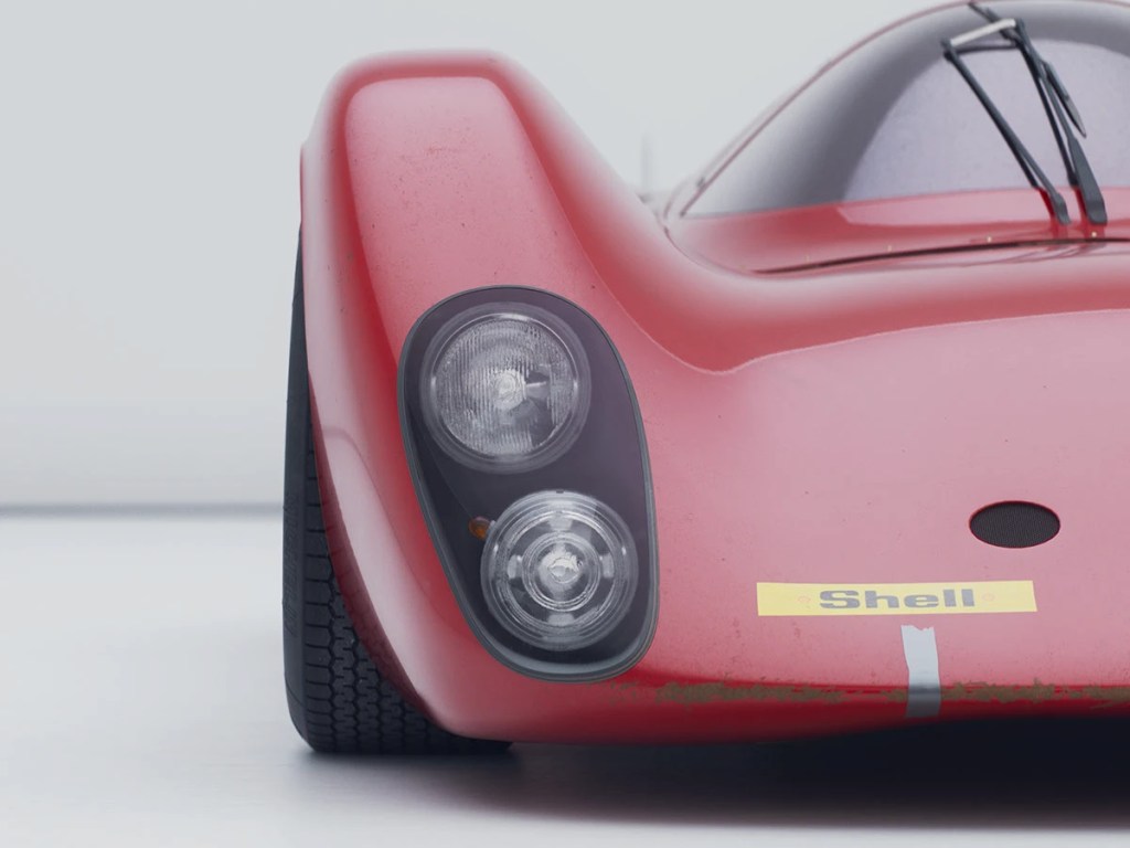 Close-up of a vintage red racing car's front, featuring round headlights and a worn texture, set against a plain background.