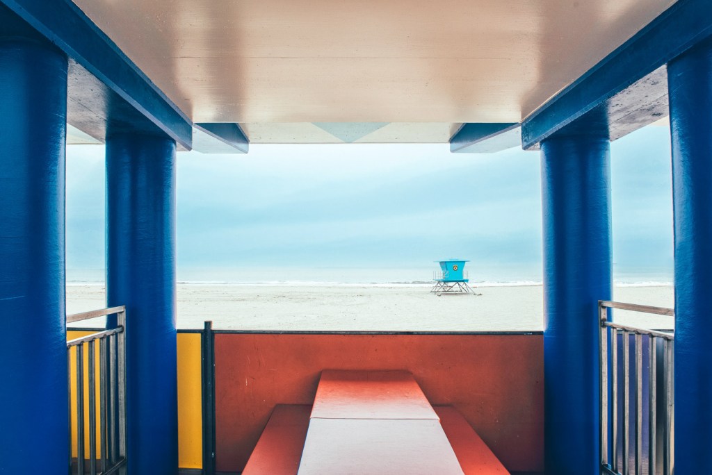 Vista desde un quiosco de playa con columnas azules y un banco rojo, mirando hacia el mar y un salvavidas azul en la distancia.