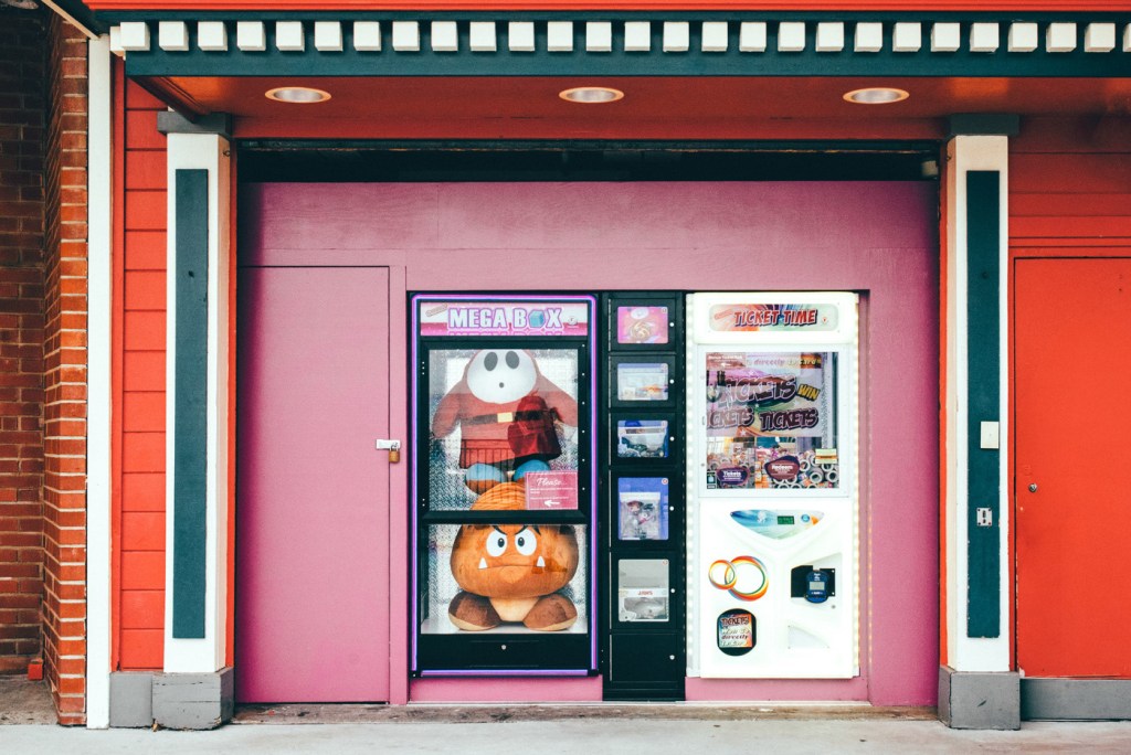 Máquina expendedora con juguetes de peluche en la parte superior, ubicada en una pared de color rosa. A la izquierda, un diseño de máquina que parece ofrecer ganancias de boletos.