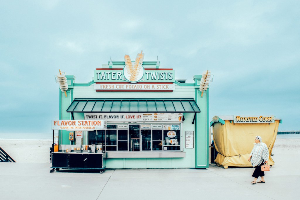 Puesto de comida llamado Tater Twists que ofrece papas fritas frescas en un palo, con una estación de sabores y una carpa de maíz asado al lado, en una playa.