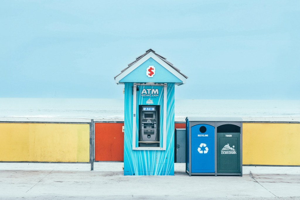Caja automática (ATM) ubicada en un paseo marítimo, junto a un recipiente de reciclaje y un basurero.