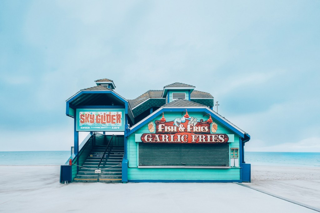 Estructura colorida de un quiosco en la playa con letreros que anuncian 'Fish & Fries' y 'Garlic Fries', y un acceso a una atracción llamada 'Sky Glider'.