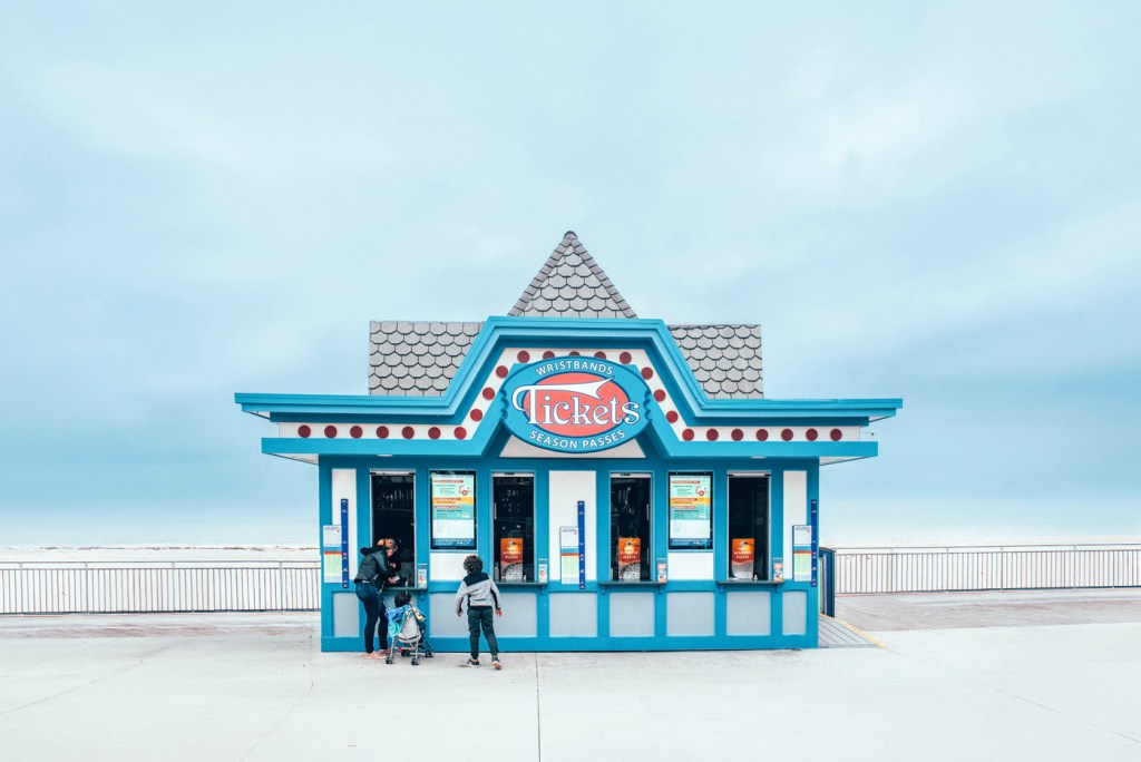 Taquilla de venta de boletos en la playa, con un diseño azul y elementos decorativos. Dos personas están de pie frente a la taquilla, una con un carrito de niño.