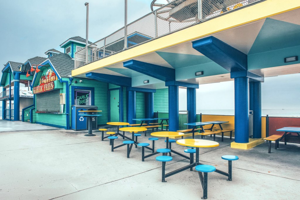 Zona de comedor al aire libre con mesas de colores amarillas y azules, junto a un restaurante de papas fritas en una estructura colorida cerca del mar.