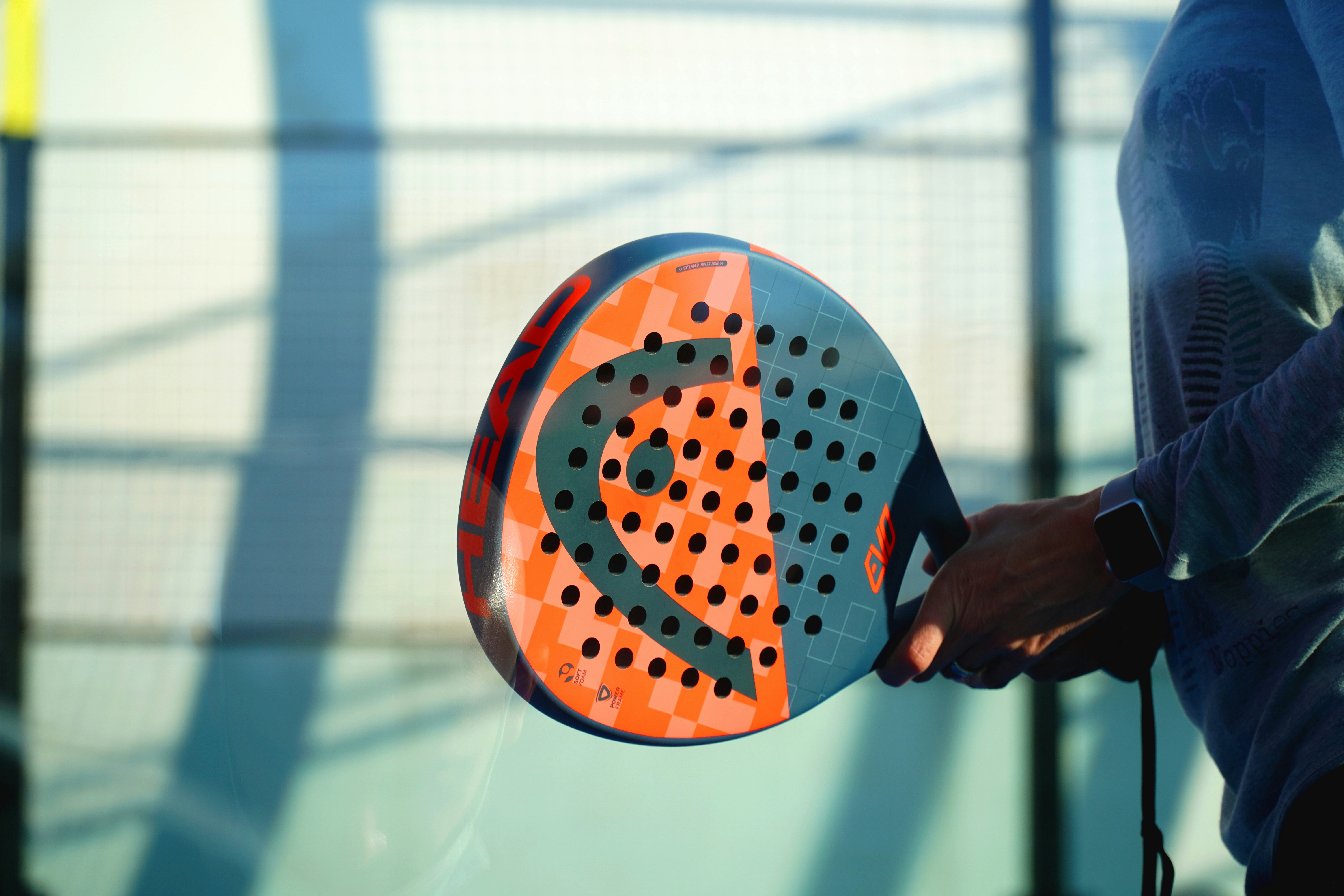 A player holds an orange and navy padel racket. The racket is round and shaped like a pickleball paddle.