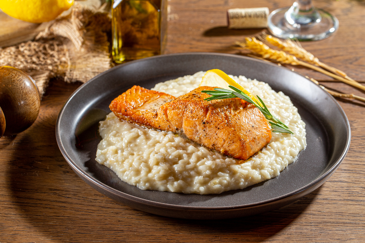 A plate of creamy risotto topped with two pieces of seared salmon, garnished with a sprig of rosemary and a lemon wedge, sits on a wooden table with olive oil and wheat in the background.