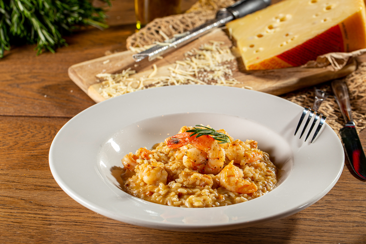 A white plate of creamy shrimp risotto garnished with rosemary sits on a wooden table. In the background, there is a wedge of cheese, a grater, and shredded cheese on a wooden board. A fork rests on the plate’s edge.