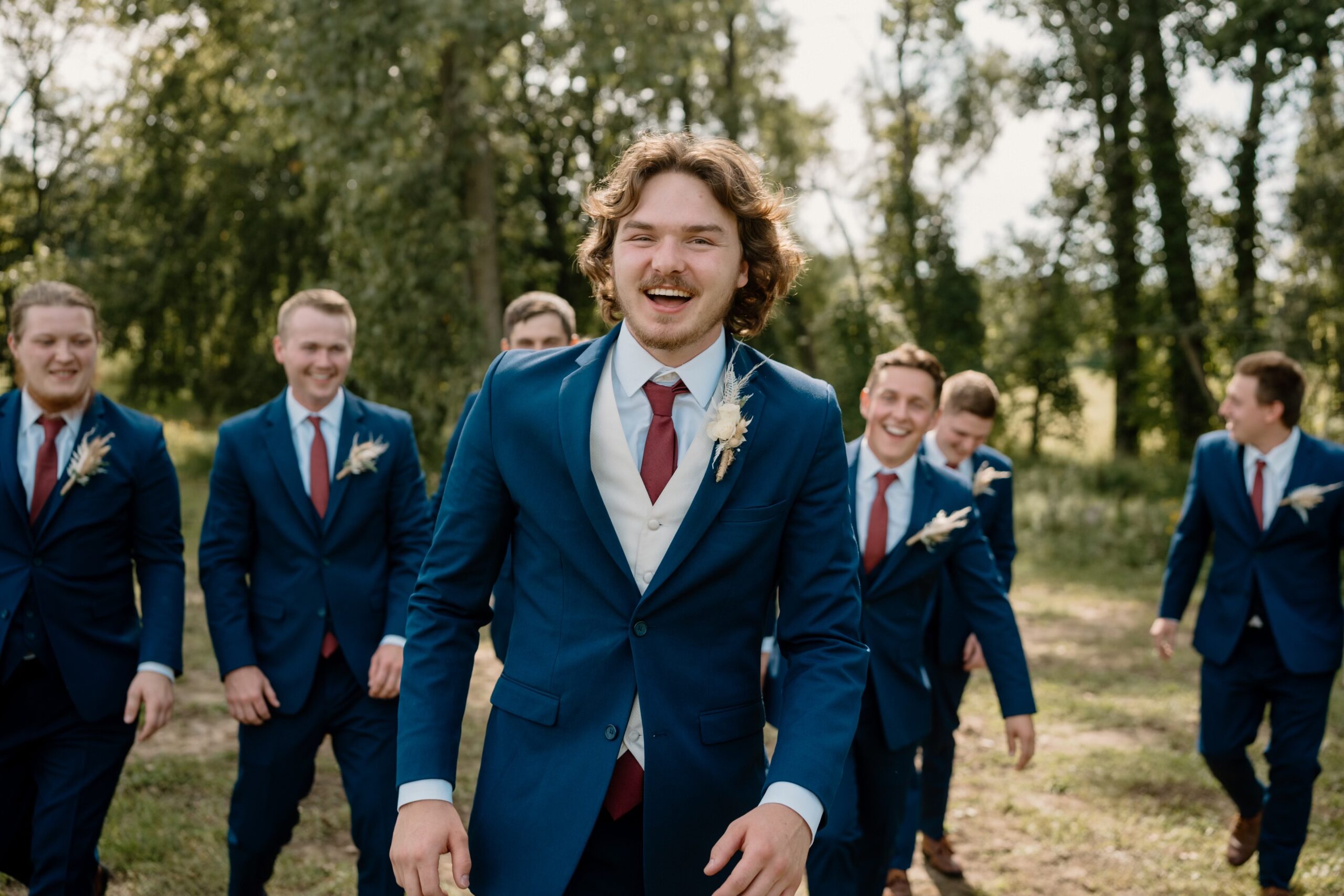 Groom and groomsmen posing outside at their wedding venue in Michigan. The groom and groomsmen are wearing cobalt and navy blue suits rented from Pierre's Bridal, Prom, and Tuxedo in Lansing, Michigan. The groom is wearing a cobalt slim fit suit, an ivory vest, and terracotta tie. The groomsmen are wearing cobalt suits, with cobalt vests, and terracotta ties. Pierre's Bridal offers a wide variety of tuxedo and suit rental and purchase options. This tuxedo shop is only about an hour and a half drive from Grand Rapids and Ann Arbor.
