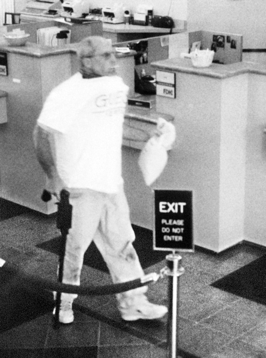 Brian Wells heads out of the PNC Bank in the Summit Towne Centre after robbing it of $8,702. Wells is carrying the cash in a white canvas bag; in his left hand is a homemade cane-shaped shotgun. He is sucking on a lollipop he grabbed while in the bank. The collar bomb is protruding from under his white T-shirt. He had demanded $250,000 from the chief teller, but left with whatever money she could give him. ERIE TIMES-NEWS, via FBI