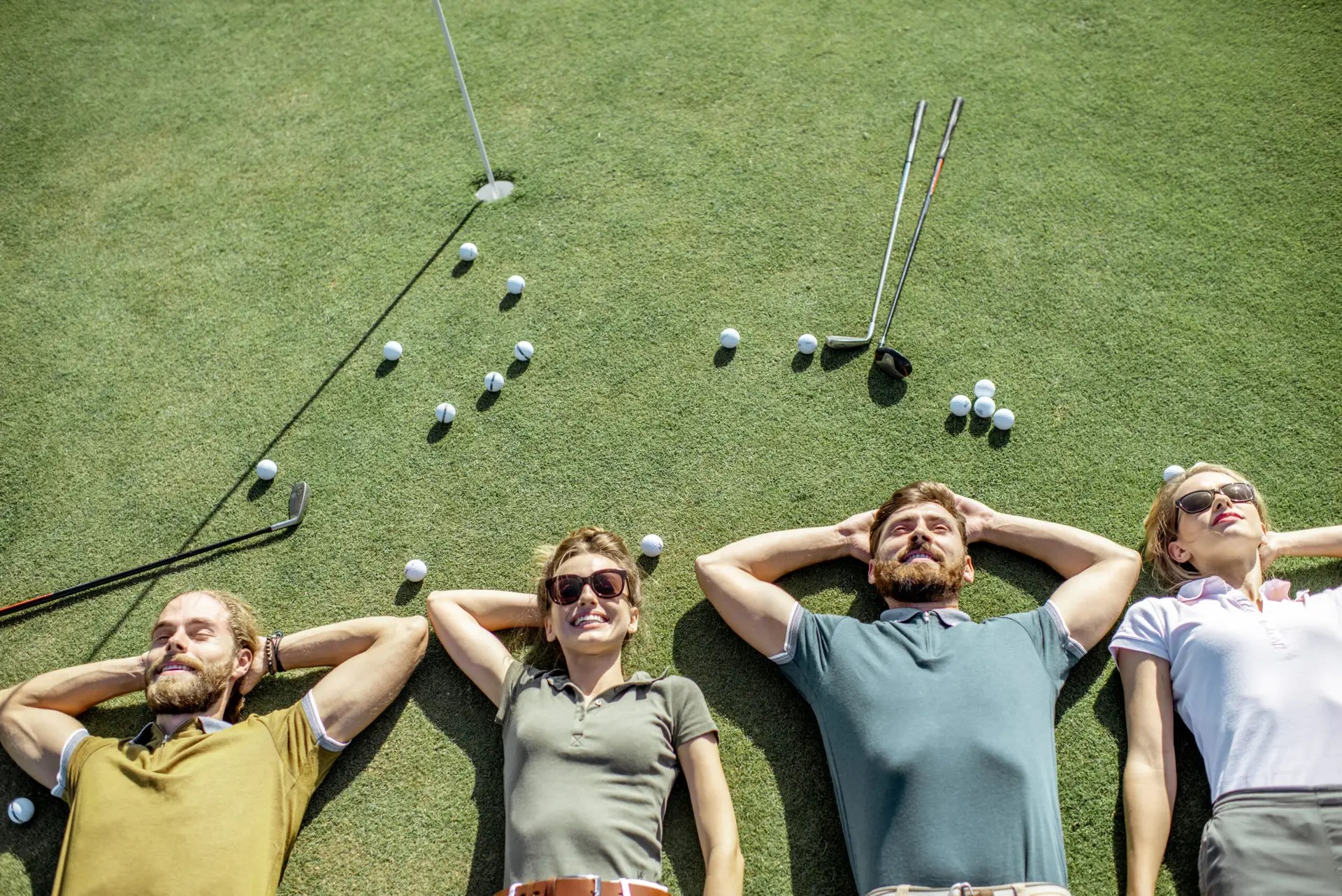 Group of a young and cheerful friends lying on the golf course with balls and putters on the grass, resting and having fun after the game