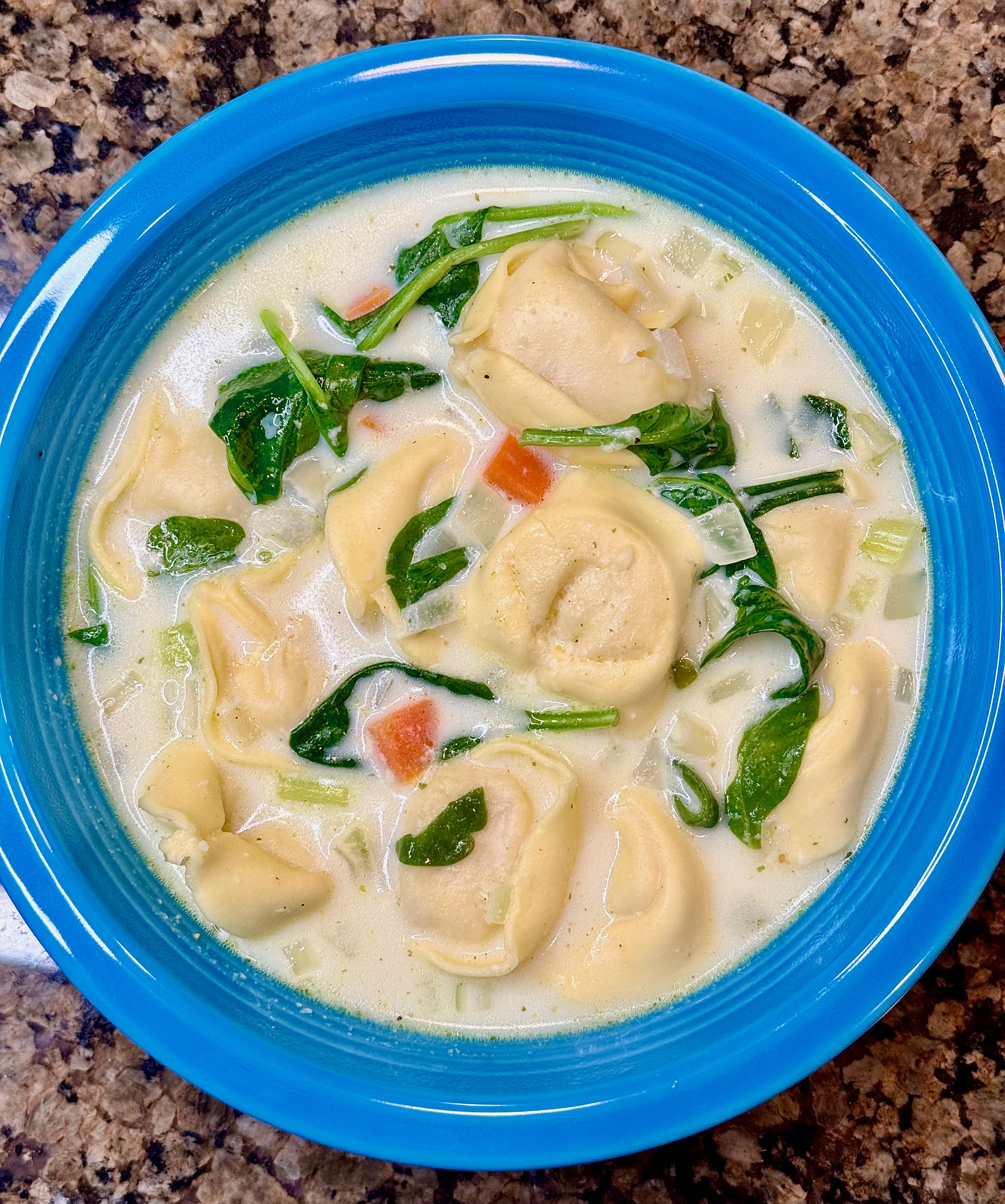 A creamy bowl of cheese tortellini soup with vegetables and herbs in a blue soup bowl