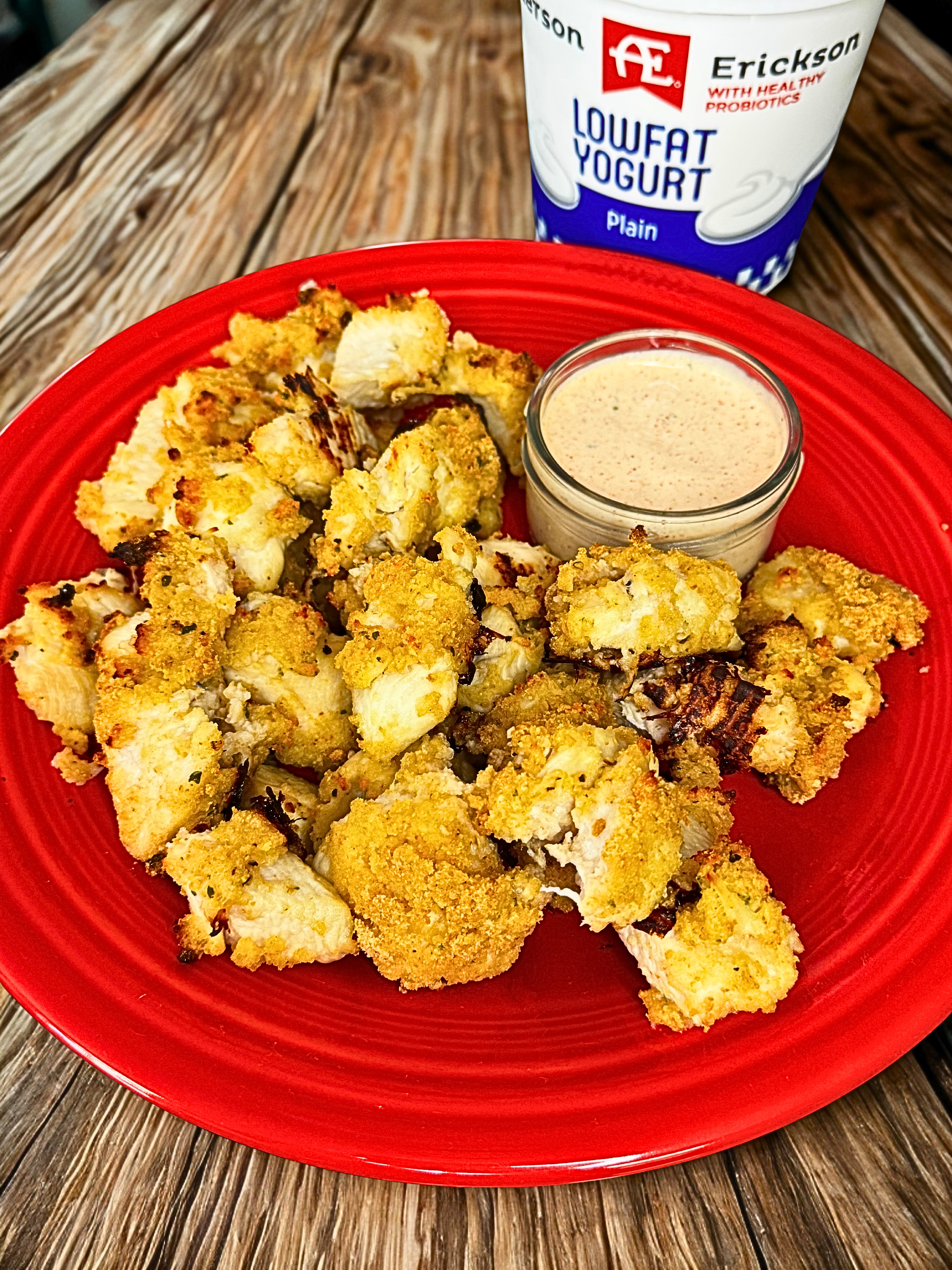 Chicken nuggets on a red plate with yogurt sitting next to it, spicy ranch in a glass jar next to it.