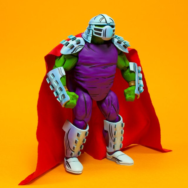 A colorful action figure depicting a muscular character with green skin, wearing a purple outfit, silver armor, and a red cape against a bright orange background.