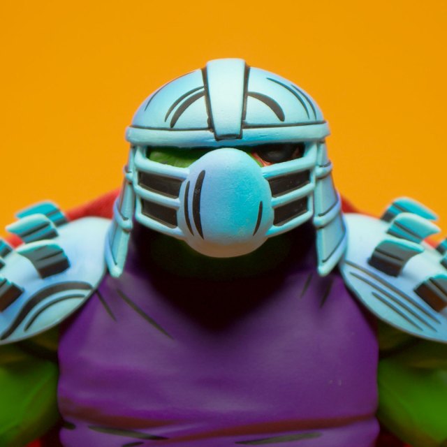 Close-up of a colorful action figure with a blue helmet and shoulder armor, wearing a purple shirt, against an orange background.