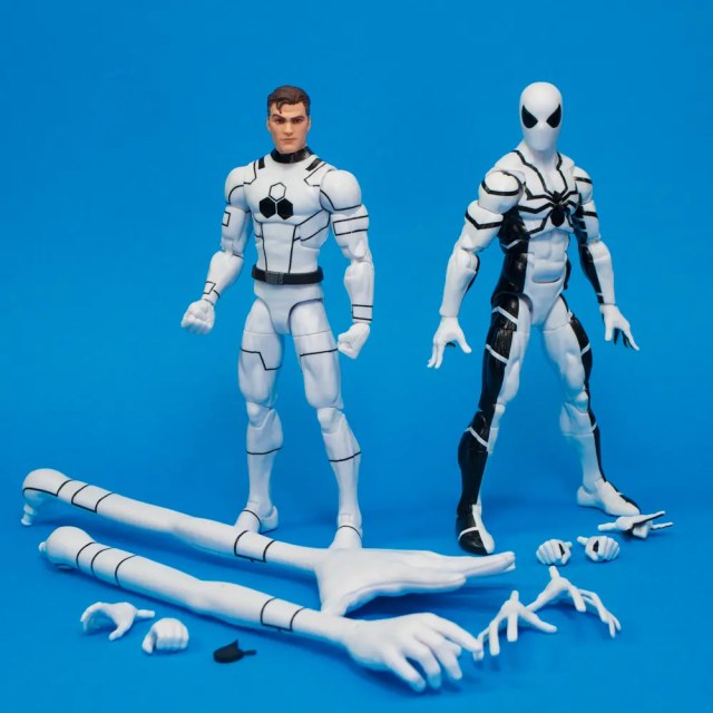 Two action figures standing on a blue background; one figure is in a white suit and the other is in a black and white spider-themed suit. Several interchangeable limbs and accessories are scattered around them.
