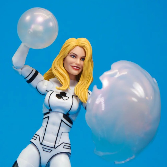 A female action figure with long blonde hair, dressed in a white and black suit, playfully holding glowing bubble-like orbs against a blue background.