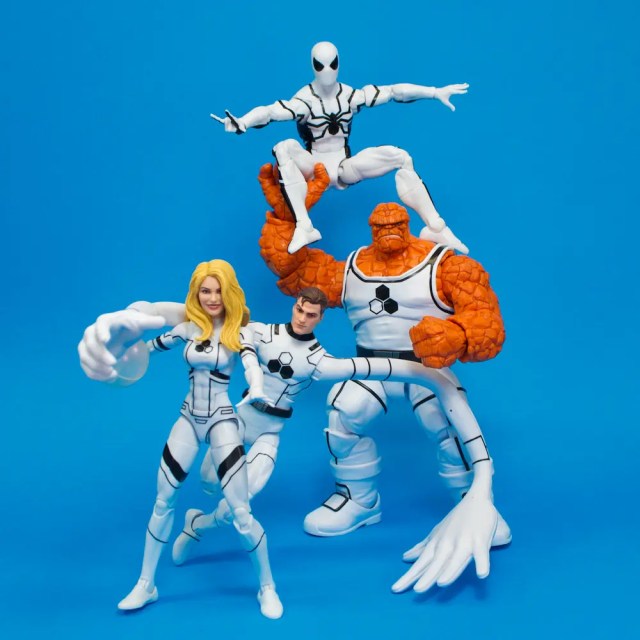 A group of four action figures in white and black costumes, featuring a female character with long blonde hair, a male character, an orange rocky figure, and a character in a black and white spider-themed suit, posed against a bright blue background.