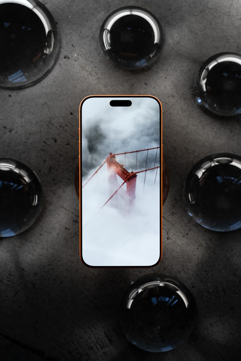 Top-down shot of an orange iPhone on a concrete surface, surrounded by large liquid drops. Oh, like liquid glass, I get it.