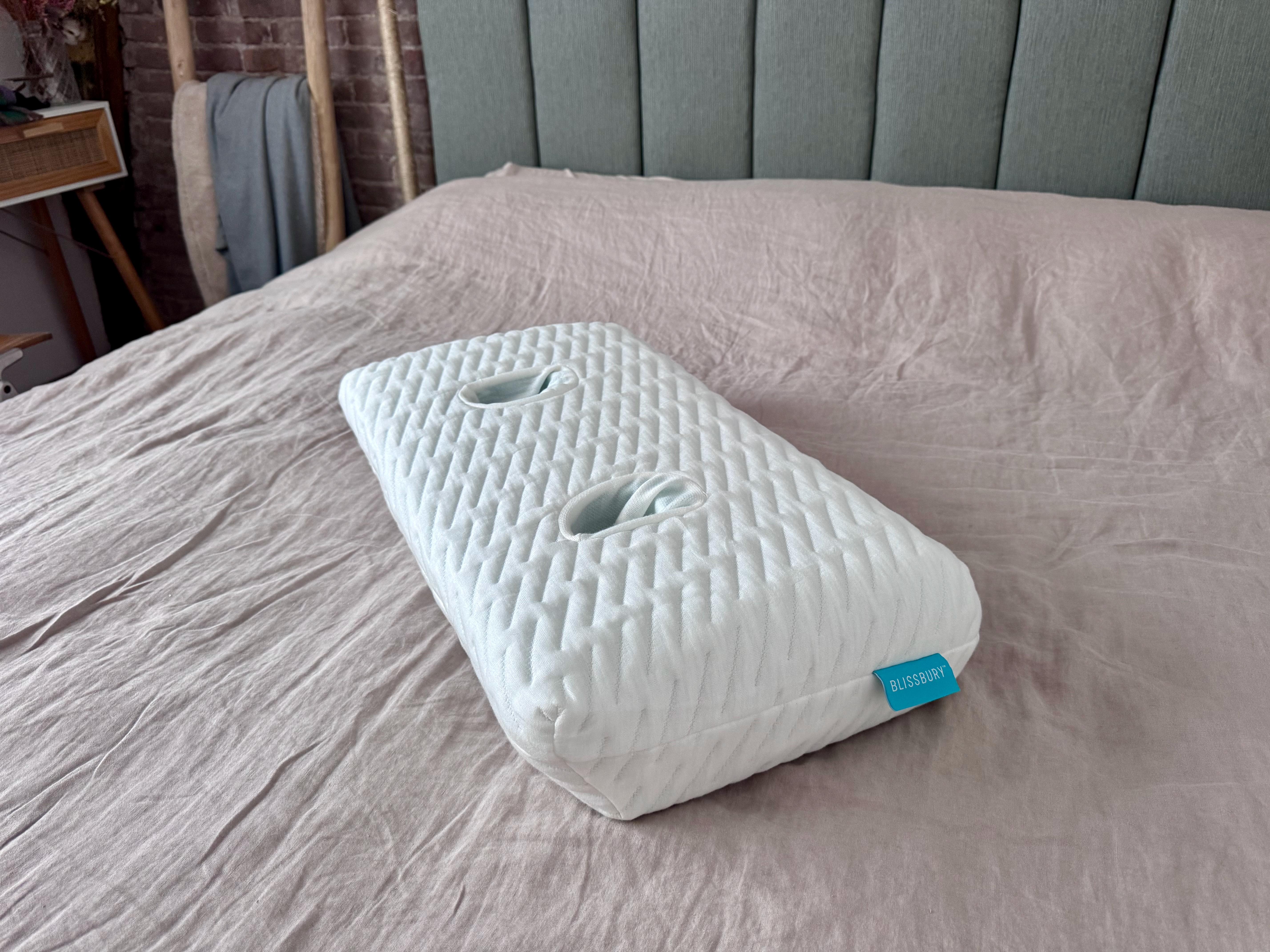 A pillow with two visible holes, equidistant from the edges on the top surface sits on a bed.