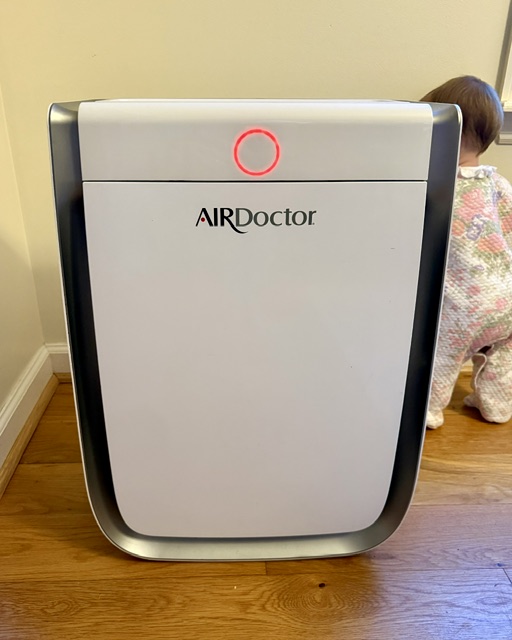 Baby standing behind AirDoctor air purifier. Air filter shows red ring, indicating poor air quality. 